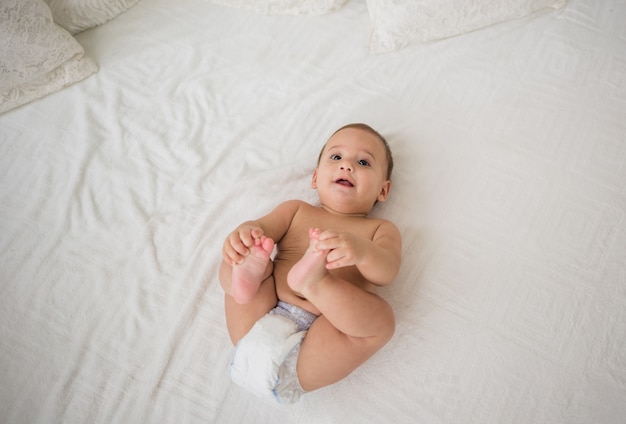 Glücklicher kleiner Junge, der in einer Windel auf einem weißen Bett liegt