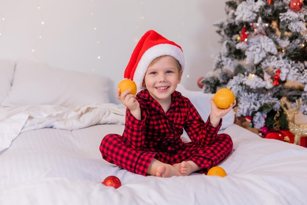 glücklicher kleiner Junge im Schlafanzug und Weihnachtsmütze zu Hause im Bett, der zu Weihnachten Mandarinen isst