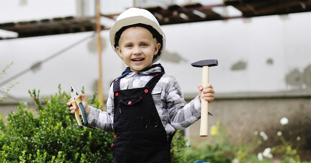 Glücklicher kleiner Junge im weißen Helm, der mit Werkzeugen auf einer Baustelle spielt Kleiner Baumeister