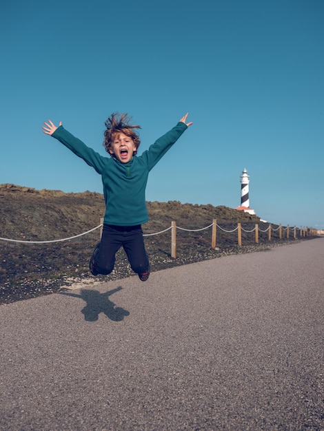 Glücklicher kleiner Junge in lässiger Kleidung, der tagsüber mit erhobenen Armen auf trockenem Weg zum Leuchtturm unter wolkenlosem blauen Himmel springt