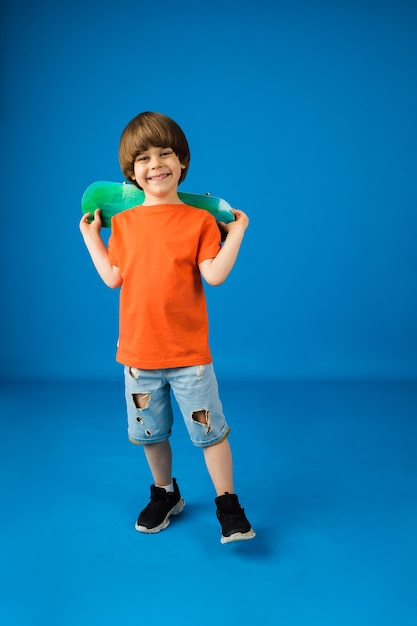 Glücklicher kleiner Junge mit braunen Haaren hält ein Skateboard auf einer blauen Oberfläche mit Platz für Text