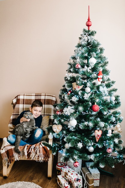 Glücklicher kleiner Junge mit Katze Weihnachtsgeschenkbox, die zu Hause in der Nähe des Weihnachtsbaums sitzt