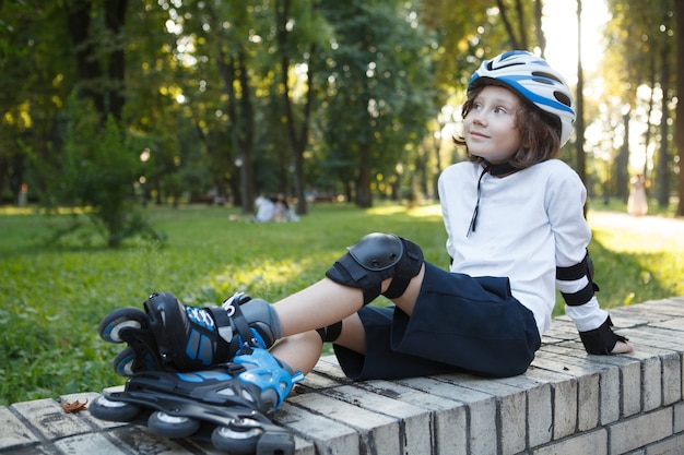 Glücklicher kleiner Junge mit Rollschuhen und Helm, der sich nach dem Skaten im Park ausruht
