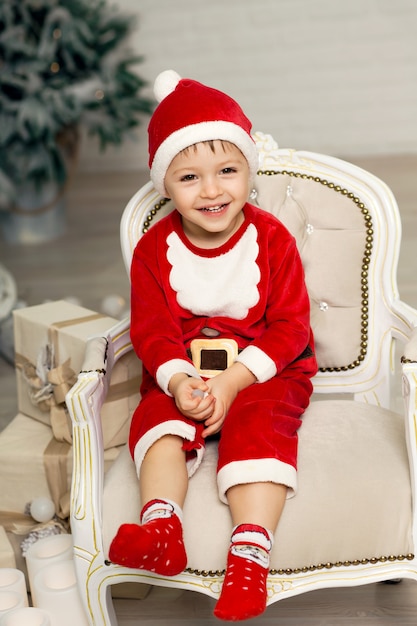 Glücklicher kleiner lächelnder Junge in Weihnachtsmann-Kostüm sitzt auf Lehnsessel nahe Weihnachtsbaum.