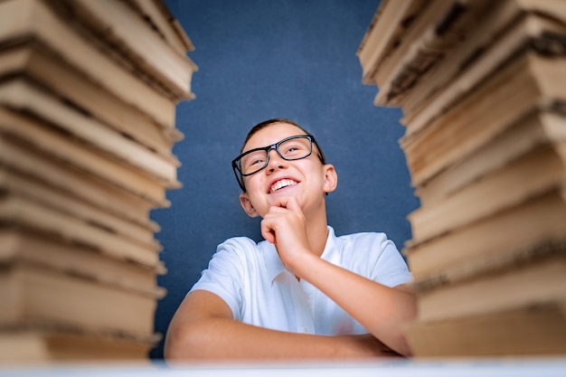 Glücklicher kluger Junge in den Gläsern, die zwischen zwei Stapel Bücher sitzen und lächelnd aufblicken.