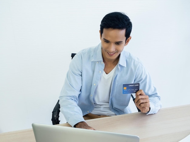 Glücklicher lächelnder asiatischer Mann im Jeanshemd, der online mit einer Kreditkarte in der Hand mit einem Laptop für sein eigenes kleines Unternehmen auf einem weißen Heimbüro-Hintergrundtisch bezahlt