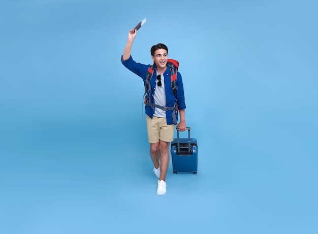 Glücklicher lächelnder asiatischer Taschenpacker-Mann mit Pass und Gepäck, die ihren Sommerferienurlaub im blauen Hintergrund genießen.