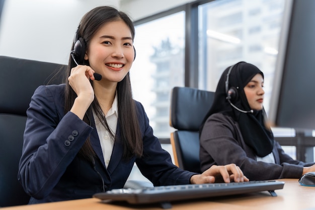 Glücklicher lächelnder betreiber asiatischer frau-kundendienstmitarbeiter mit headsets, die am computer in einem callcenter arbeiten und mit dem kunden sprechen, um zu helfen, das problem mit ihrem service-verstand zu lösen