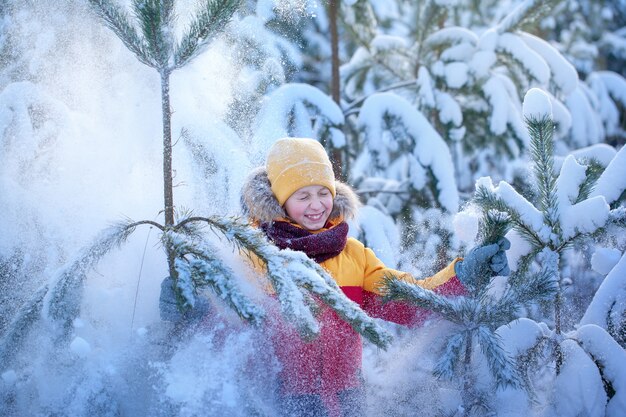 Glücklicher lächelnder Junge in Winterkleidung, der im verschneiten Winterwald spaziert. Kinder spielen draußen im Schnee.