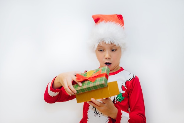Glücklicher lächelnder kaukasischer Junge im Sankt-T-Shirt und im Hut, der eine Pocken mit Weihnachtsgeschenk öffnet. Foto in hoher Qualität