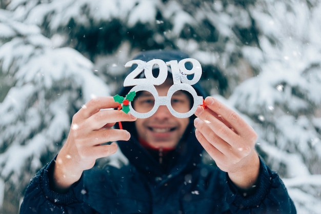 Glücklicher lächelnder Mann, der Gläser des neuen Jahres der Partei mit Nr. 2019 im Winterpark hält