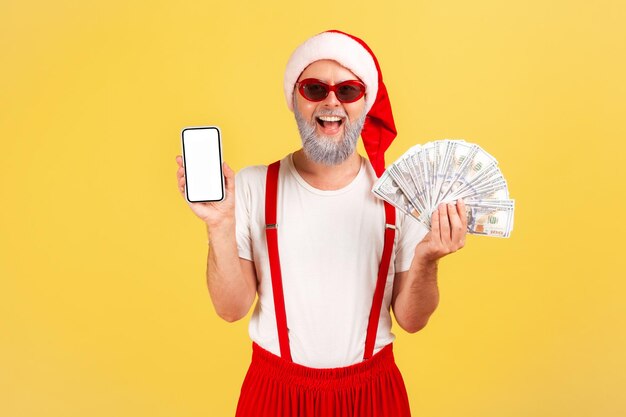 Glücklicher lächelnder Mann im Weihnachtsmann-Kostüm und stylische rote Sonnenbrille, die Dollarfächer hält und Smartphone mit leerem Bildschirm zeigt, Wett-App. Studioaufnahme im Innenbereich isoliert auf gelbem Hintergrund