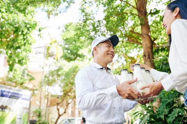 Glücklicher lächelnder vietnamesischer Lieferbote, der der hübschen jungen Frau Satz Milchflaschen gibt