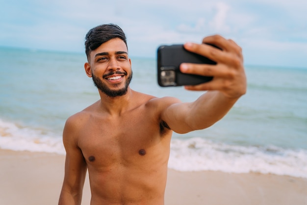 Glücklicher lateinamerikanischer Mann, der selfie Strandhintergrund, sonnige Sommerfarben macht