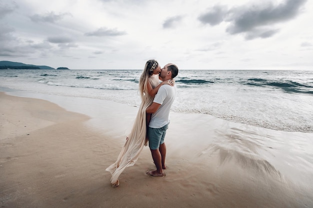 Glücklicher, liebevoller Ehemann und Ehefrau, gekleidet in Weiß, küssen sich an einem exotischen Strand, der Mann umarmt das Mädchen, während er sie vom Boden hebt.