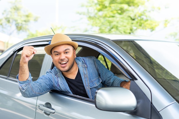 Glücklicher männlicher Fahrer, der lächelt, während er in einem Auto mit offener Frontscheibe sitzt. Junger asiatischer Mann lächelt und schaut durch das Fenster und genießt die Reise