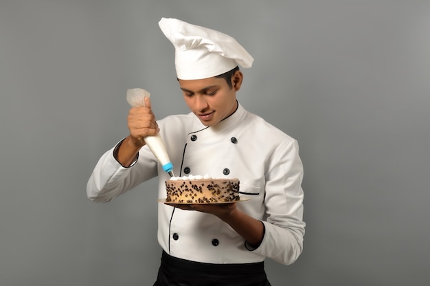 Glücklicher männlicher indischer Koch Konditor, der Schokoladenkuchen dekoriert, der das Sahnehäubchen auf dem Kuchen macht
