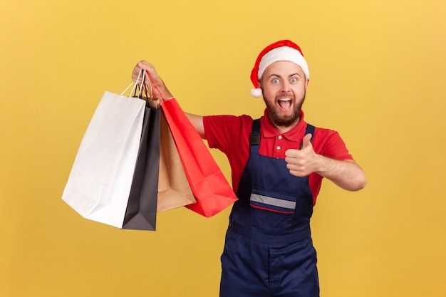 Glücklicher männlicher Kurier in Uniform und Weihnachtsmann-Hut, der Daumen nach oben zeigt und Papiereinkaufstüten hält