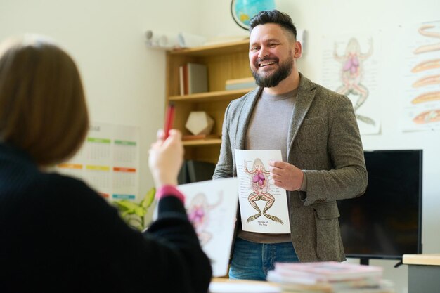 Glücklicher männlicher Lehrer mit Poster der inneren Struktur des Frosches, der den Schüler anschaut