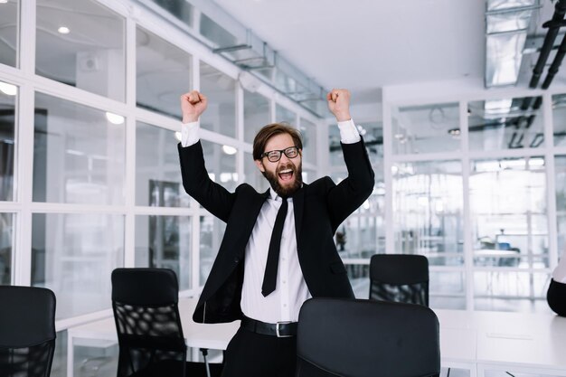 Glücklicher männlicher Manager im schwarzen Anzug mit Bart und Brille feiert seinen Erfolg, indem er im Büro neben dem Tisch tanzt