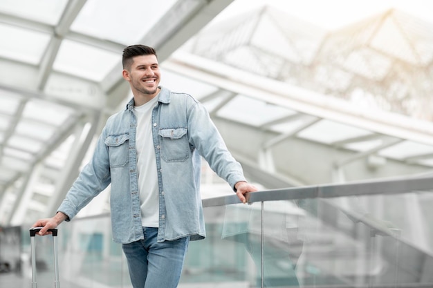 Glücklicher männlicher Tourist, der mit Koffer im modernen Flughafen steht