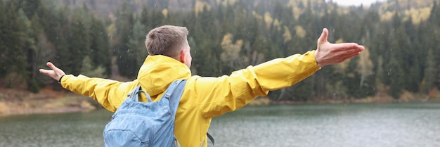 Glücklicher männlicher Tourist mit offenen Armen steht am Ufer des Sees und blickt auf die Berge im Freien