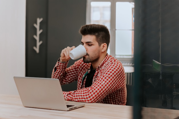 Glücklicher Mann, der an Laptop arbeitet und zu Hause Kaffee trinkt.