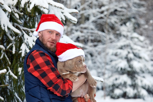 Glücklicher Mann, der einen schönen Hund in seinen Händen hält, der in einer Weihnachtsmütze im verschneiten Wald trägt Lächelnder Junge, der im Winterholz einen entzückenden Welpen umarmt Haustierliebhaber Konzept des Freundes des Hundemenschen Weihnachtsferien