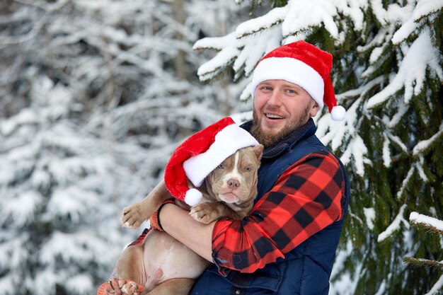 Glücklicher Mann, der einen schönen Hund in seinen Händen hält und eine Weihnachtsmütze im verschneiten Wald trägt. Lächelnder Junge, der entzückenden Welpen im Winterholz umarmt. Haustier Liebhaber. Hund - Freundkonzept des Menschen. Weihnachtsferien