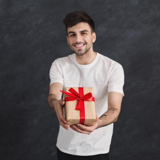 Glücklicher Mann, der Geschenk mit rotem Bandbogen in seinen Händen auf schwarzem Studiohintergrund hält. Weihnachten oder Valentinstag, Urlaubskonzept, Kopierraum