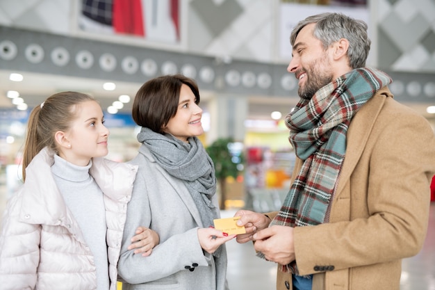 Glücklicher Mann, der seiner Frau und Tochter Kreditkarte gibt, bevor er am schwarzen Freitag oder am Weihnachtsverkauf im Einkaufszentrum einkauft