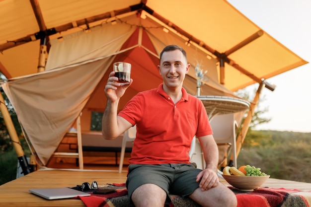 Glücklicher Mann, der sich am Sommerabend beim Glamping entspannt und Wiskiey mit Cola trinkt Luxus-Campingzelt für Erholung im Freien und Erholung Lifestyle-Konzept