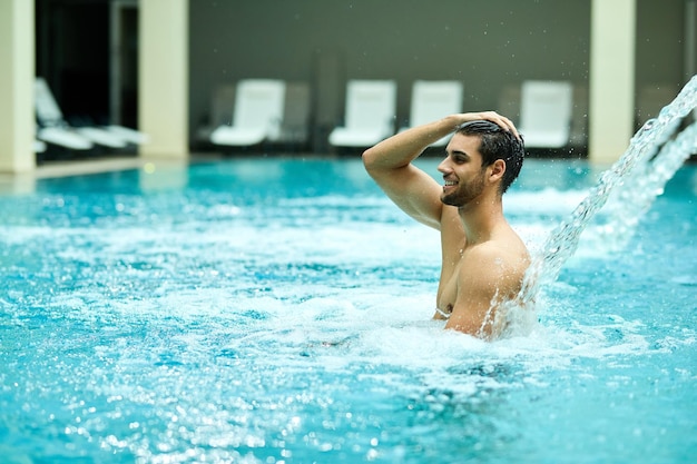 Glücklicher Mann, der sich unter einem Wasserstrahl im Schwimmbad entspannt