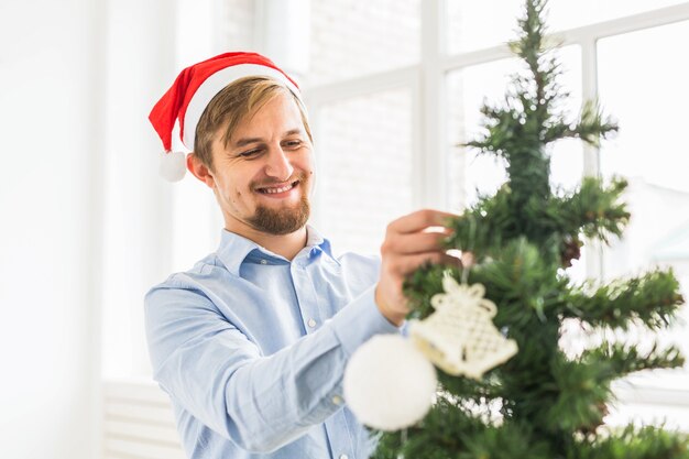 Glücklicher Mann, der zu Hause Weihnachtsbaum mit Weihnachtsmann-Hut schmückt. Mann, der während der Winterferien Baum mit Kugeln schmückt.