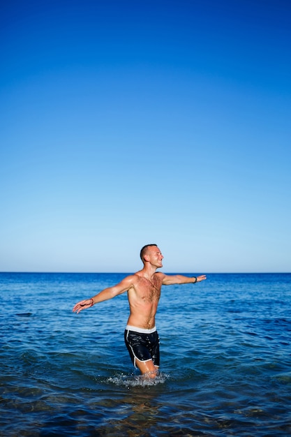 Glücklicher Mann Freude im Urlaub, steht auf See und spritzt im Wasser. Herrenshorts, Sonne, Strand