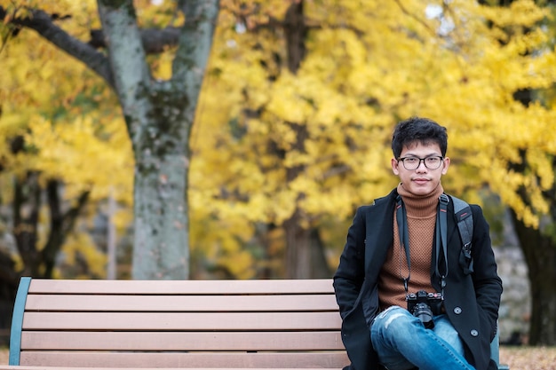 Glücklicher Mann genießt im Park im Freien in der Herbstsaison Asiatischer Reisender in Mantel und Kamera vor gelbem Ginkgo-Blätter-Hintergrund