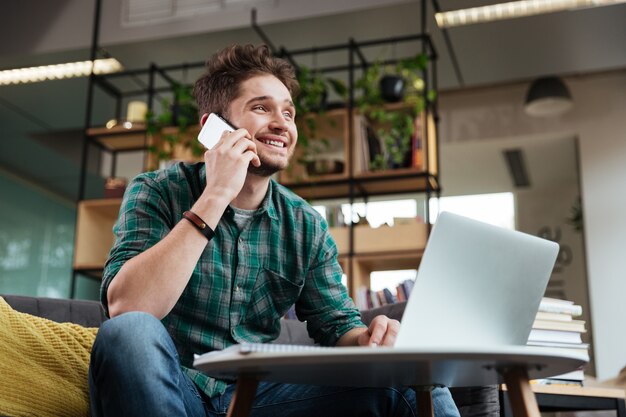 Glücklicher Mann im grünen Hemd, der mit Laptop auf dem Sofa am Tisch sitzt und im Büro telefoniert.