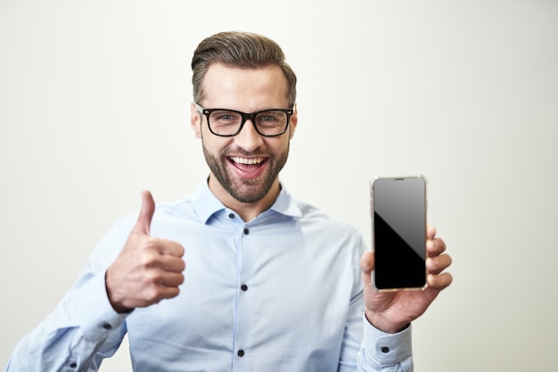 Glücklicher Mann in Hemd und Brille, der seinen Daumen hochlegt und den Smartphone-Bildschirm zeigt. Raumfoto kopieren