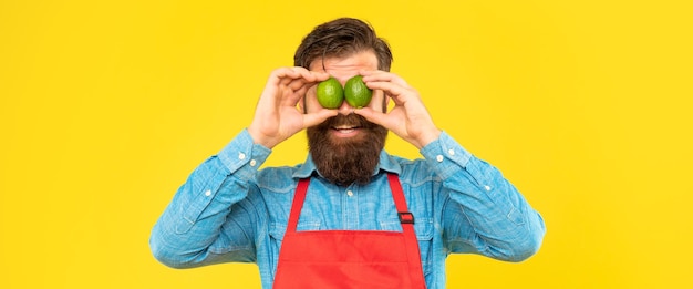 Glücklicher Mann in Schürze, der frische Limetten auf den Augen hält, gelber Hintergrund Obstverkäufer