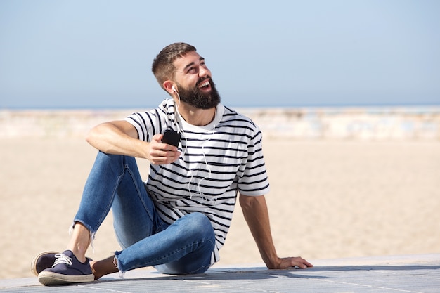 Glücklicher Mann in voller Länge in den Kopfhörern, die auf Strand mit Handy sitzen