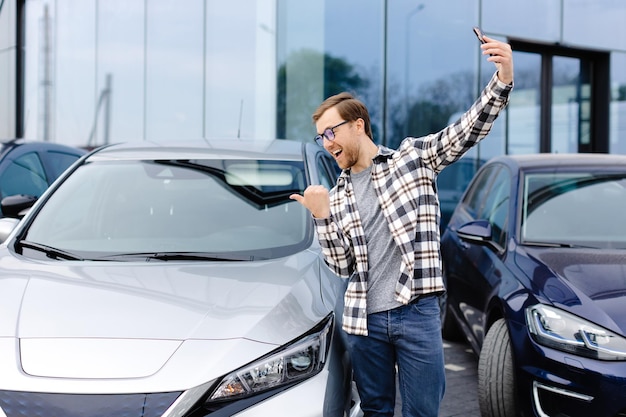 Glücklicher Mann macht nach dem Kauf eines neuen Autos ein Selfie mit dem Smartphone. Der Mann zeigt das gekaufte Auto per Videolink am Telefon