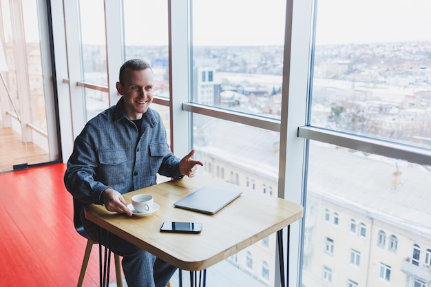 Glücklicher Mann mit Kaffee, der per Smartphone anruft, während er am Schreibtisch einen Laptop mit Fernzugriff verwendet. Fröhlicher Freiberufler, der eine Kaffeepause genießt, während er aus der Ferne arbeitet