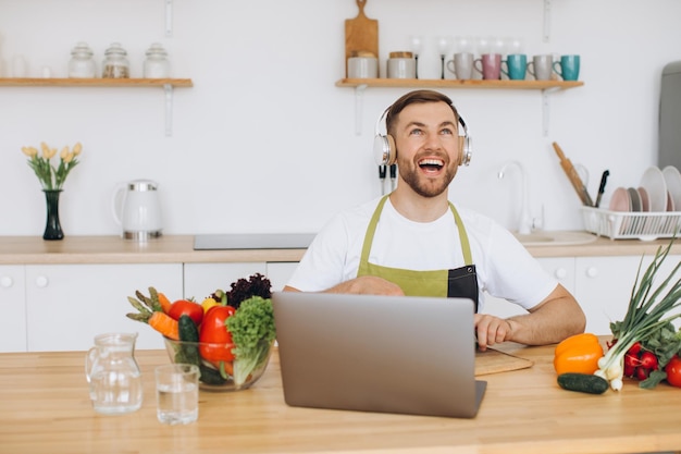 Glücklicher Mann mit Kopfhörern bereitet in der Küche Salat aus frischem Gemüse zu und schaut auf den Laptop