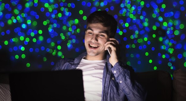 Glücklicher mann mit laptop und smartphone nachts. junger mann, der spät im homeoffice am bokeh-hintergrund arbeitet, kopienraum