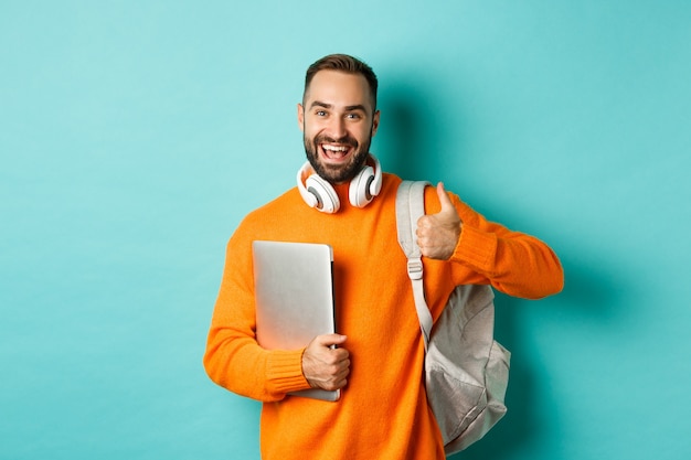 Glücklicher mann mit rucksack und kopfhörern, der laptop hält und lächelt, daumen hoch zur zustimmung zeigend, über türkisfarbenem hintergrund stehend