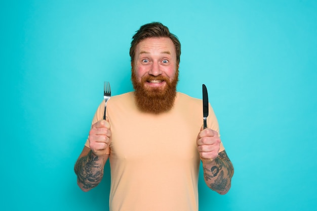 Glücklicher Mann mit Tattoos ist bereit, mit Besteck in der Hand zu essen