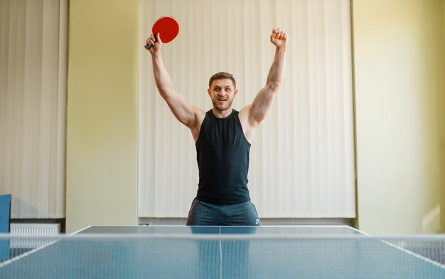 Glücklicher Mann mit Tischtennisschläger hob die Hände, Turnier drinnen. Männliche Person in Sportbekleidung, Training im Tischtennisclub