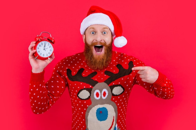 Foto glücklicher mann mit weihnachtsmann-hut, der mit dem finger auf die weckerzeit des neujahrs-countdowns zeigt