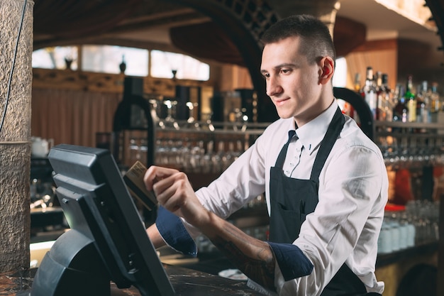 glücklicher Mann oder Kellner in der Schürze am Schalter mit Kasse, die an der Bar oder im Café arbeitet