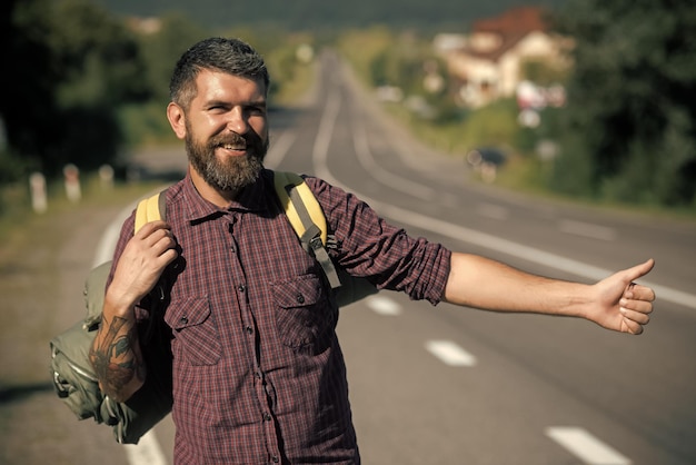 Glücklicher Mann Sommerurlaub Konzept Mann mit Rucksack glücklich Trampen auf der Straße Abenteuer Entdeckung Fernweh Hipster Wanderer zeigen Daumen nach oben Handgeste an einem sonnigen Tag Tourist Traveller Travel Auto Stop